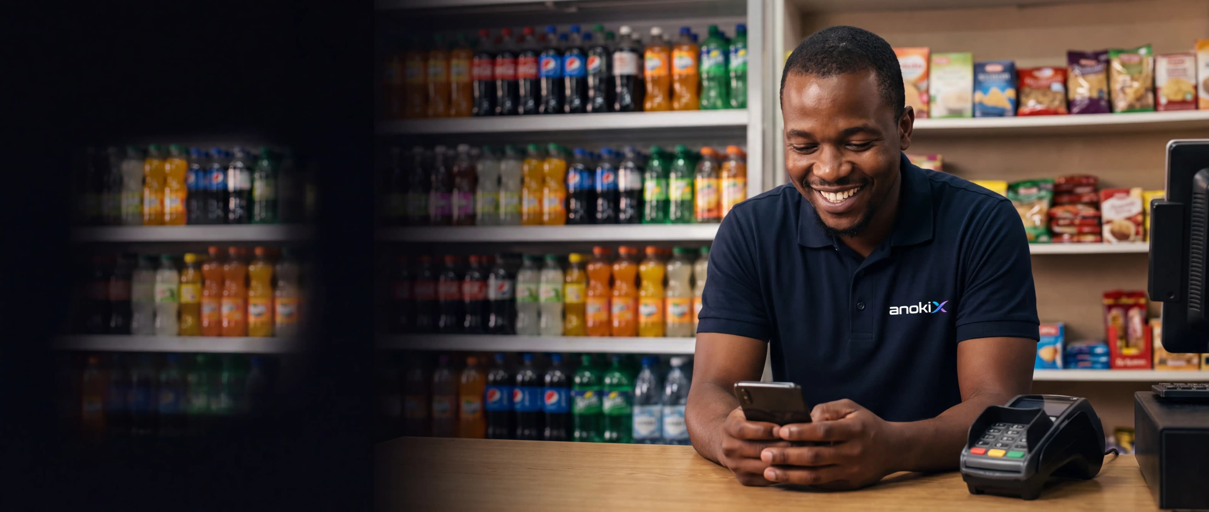 Shop owner smiling at his phone behind the counter of his grocery shop, a card payment terminal beside him